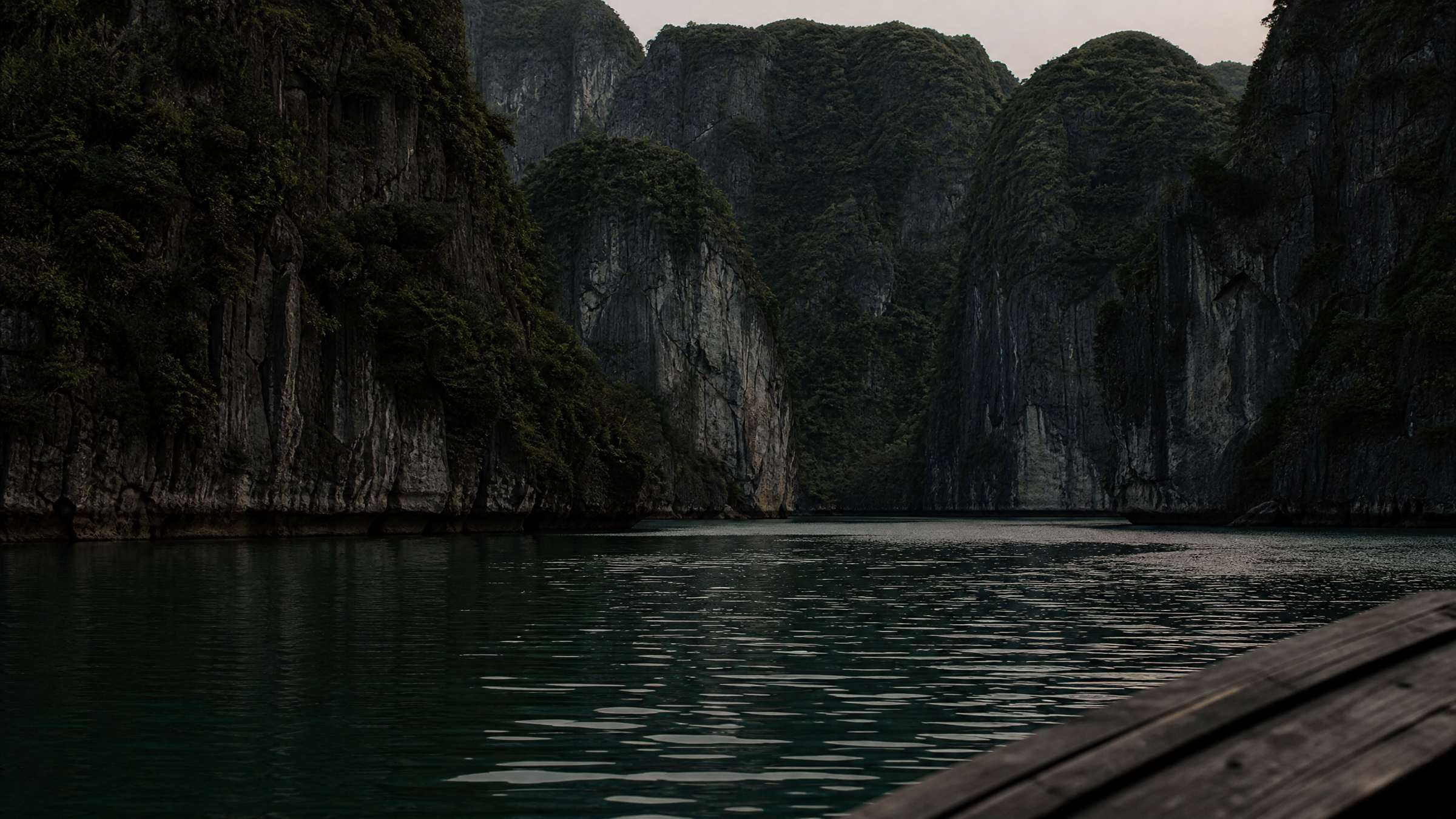 A bay of limestone karst islands from the deck of a small wooden boat, low to deep jade water in the late afternoon.