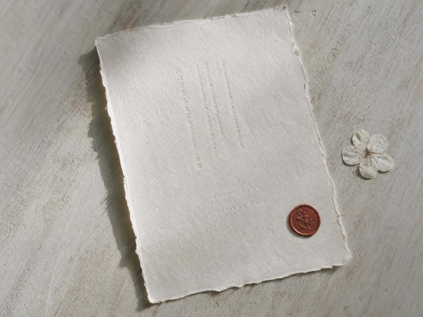 The printed invitation lying on pale wood: a single sheet with the couple's names in mincho, a small red seal, and one pressed blossom beside it.