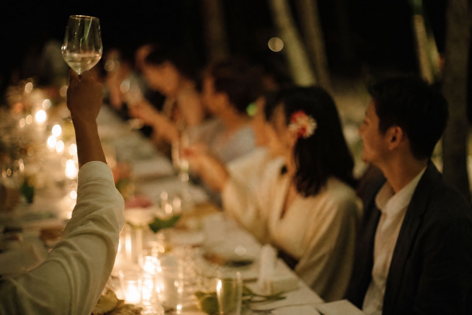 A hand raised mid-toast at the end of the long table after dark, the rest of the table soft behind it in lantern light.