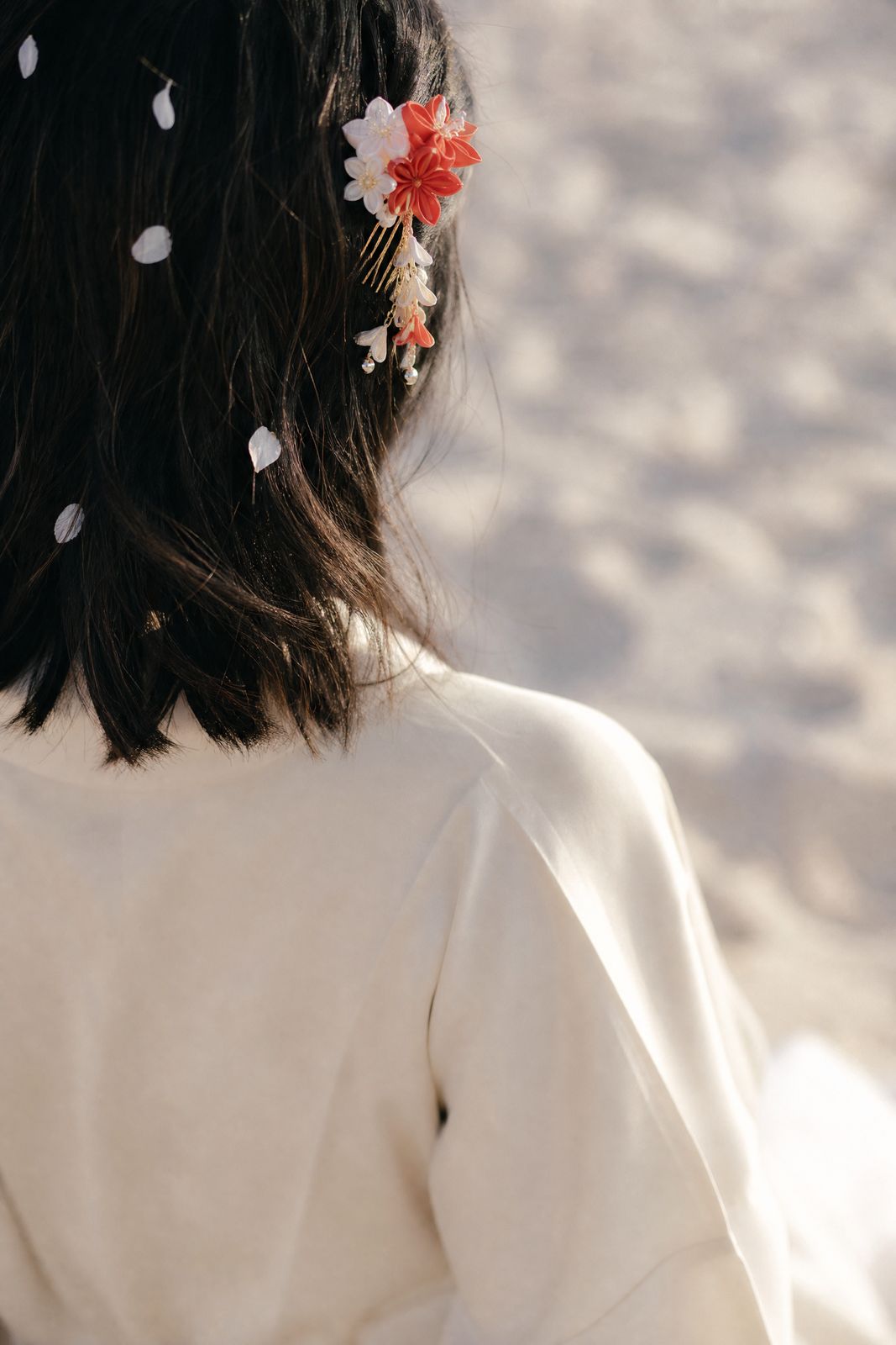Hana photographed from behind, the red-and-white kanzashi comb above her ear and white petals caught in dark hair.
