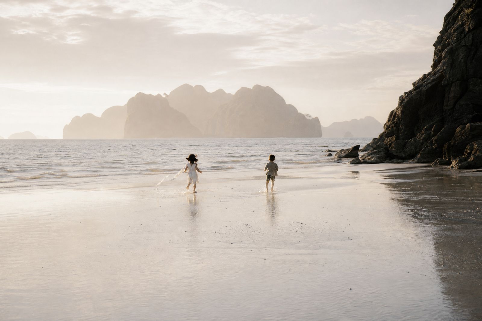 Two children running along the wet tideline in the last of the light, karst islands in warm haze behind them.