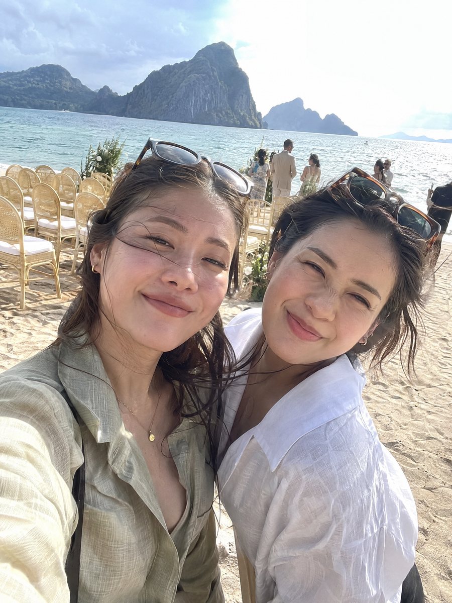 Two friends squinting into a beach selfie, a karst island behind them.