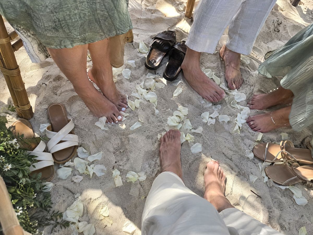 A row of bare feet and kicked-off sandals in the sand, petals between them.