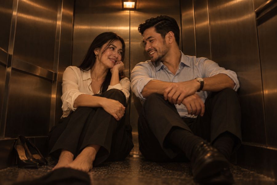 Nadine and Rafael sitting on the floor of a stalled lift, lit only by its amber emergency lamp, laughing as they talk.