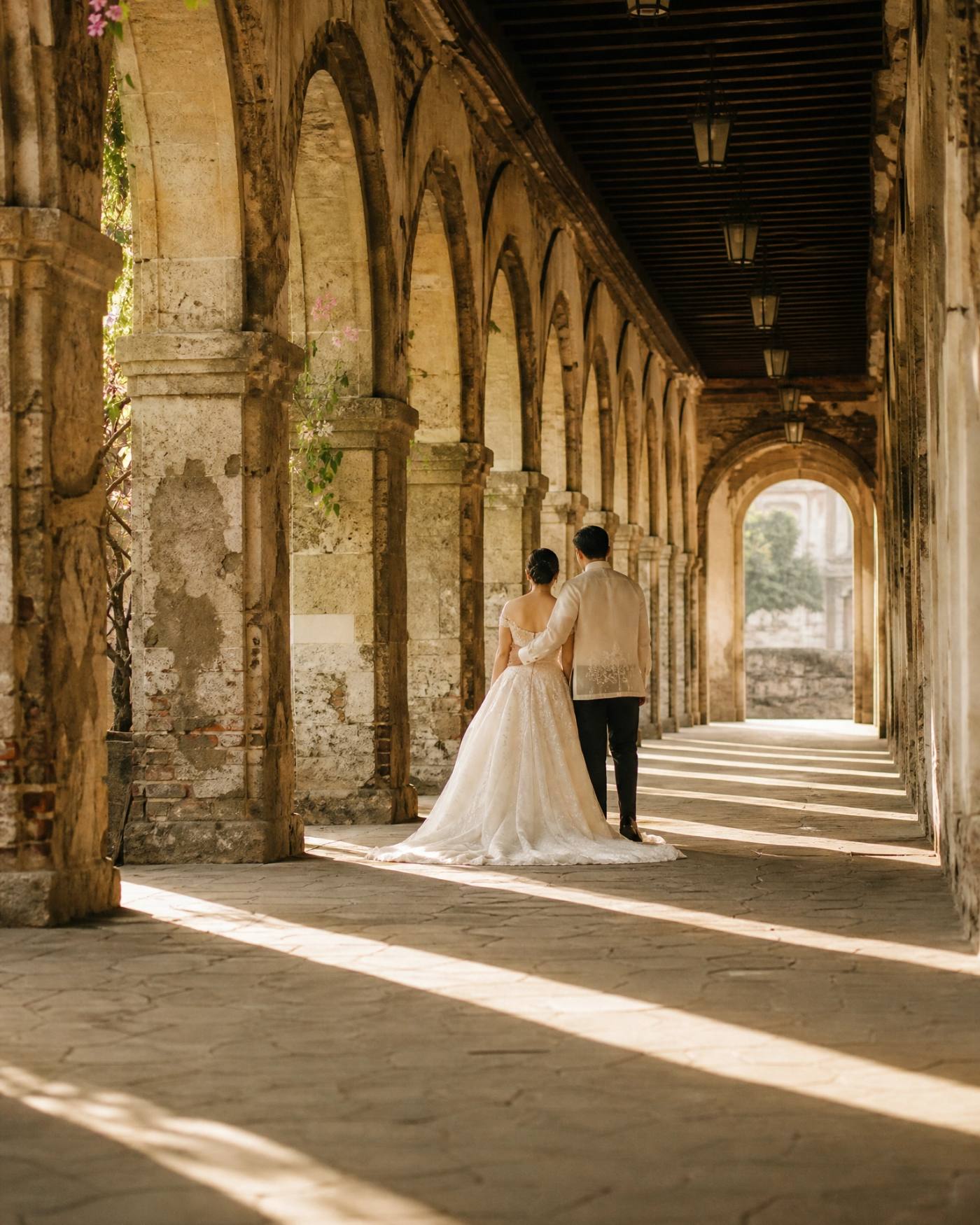 Andrea and Paolo standing together in a stone arcade, she in a long ivory gown seen from behind, he in an embroidered pi&ntilde;a barong.