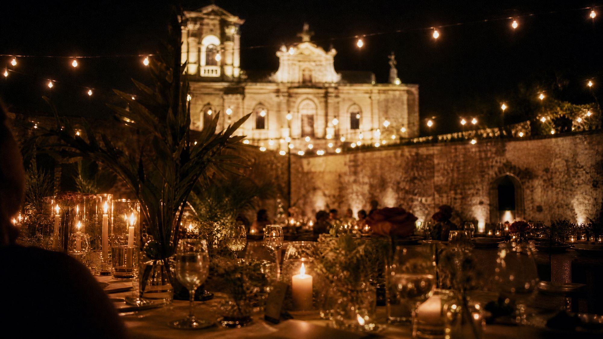 The courtyard at night: candles and glasses down a long table, festoon lights overhead and the lit parish fa&ccedil;ade beyond the wall.