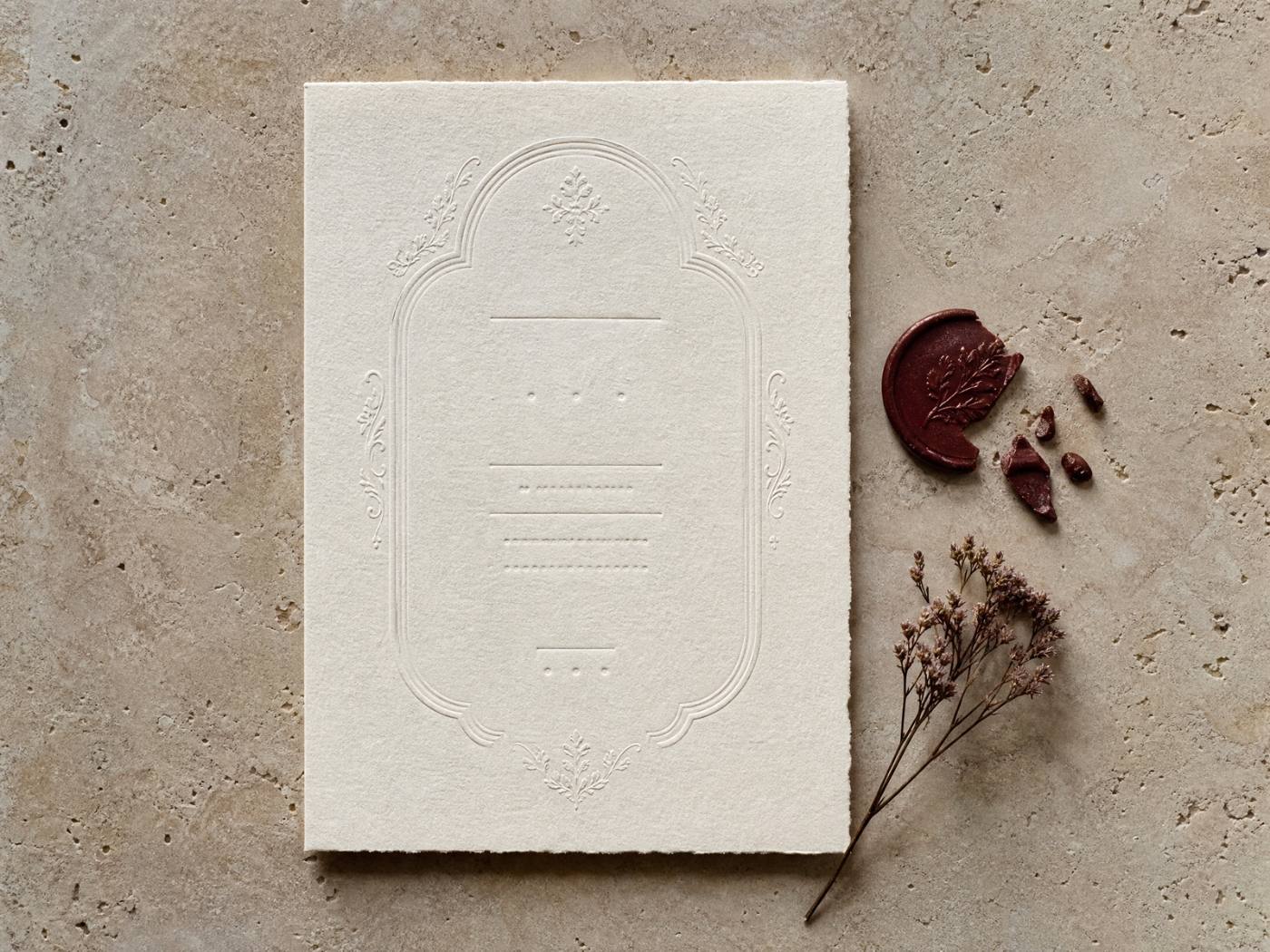 The printed invitation lying on pale stone: a deckle-edged sheet of ivory paper blind-embossed with a scalloped frame, the broken burgundy wax seal and three chips of it beside the sheet, and a dried sprig laid below.