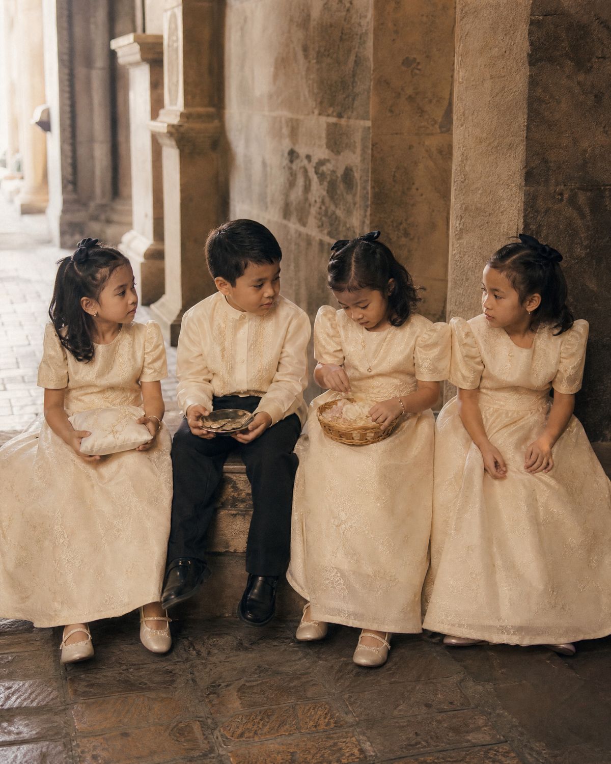 Four small children in ivory Filipiniana sitting in a row on a low stone bench, holding a ring pillow, a dish of coins and a basket of petals.
