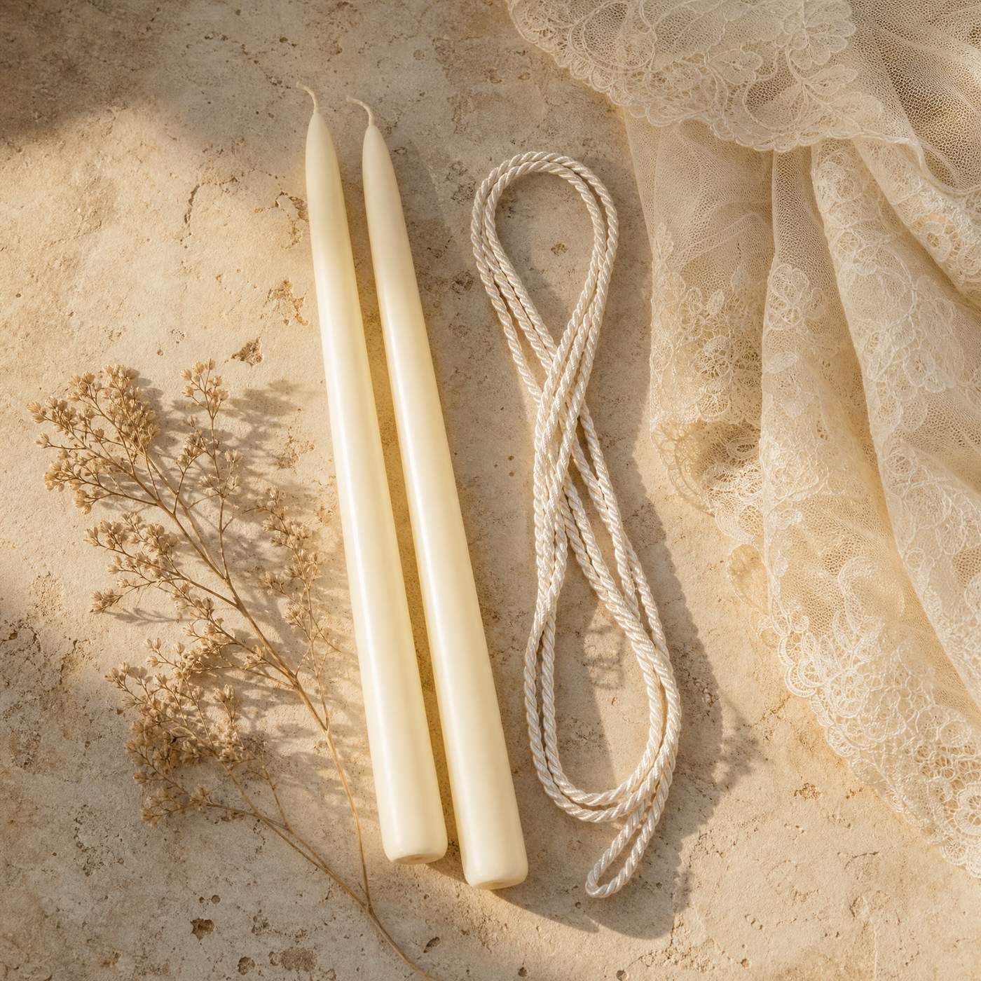 Two unlit ivory candles, a looped white silk cord and a folded lace veil laid out on pale weathered adobe stone.