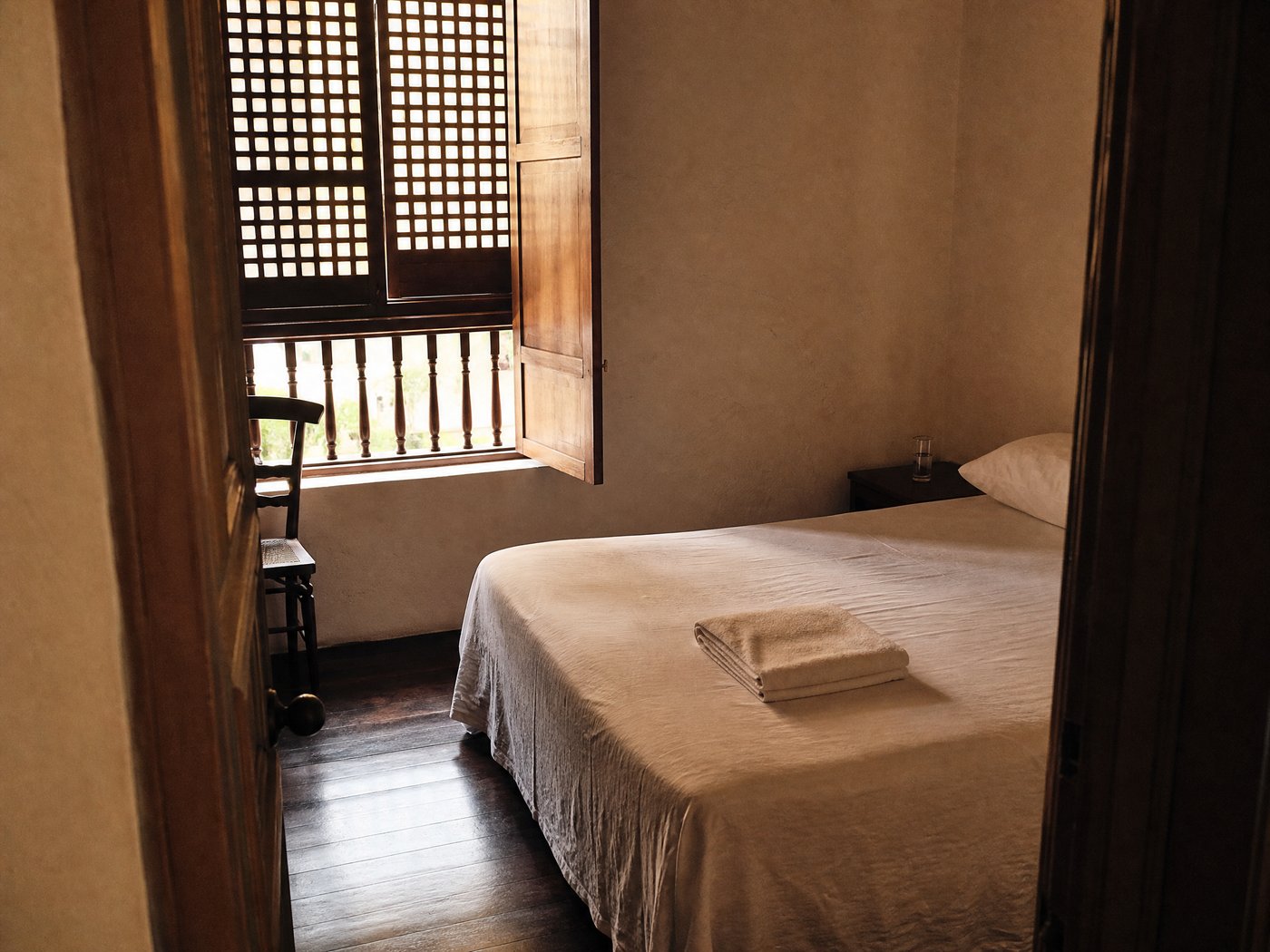 A small guest room seen through an open doorway: a low bed under a plain cream cover with a folded towel on it, a dark wooden floor, and a shuttered window standing open onto a balustrade.