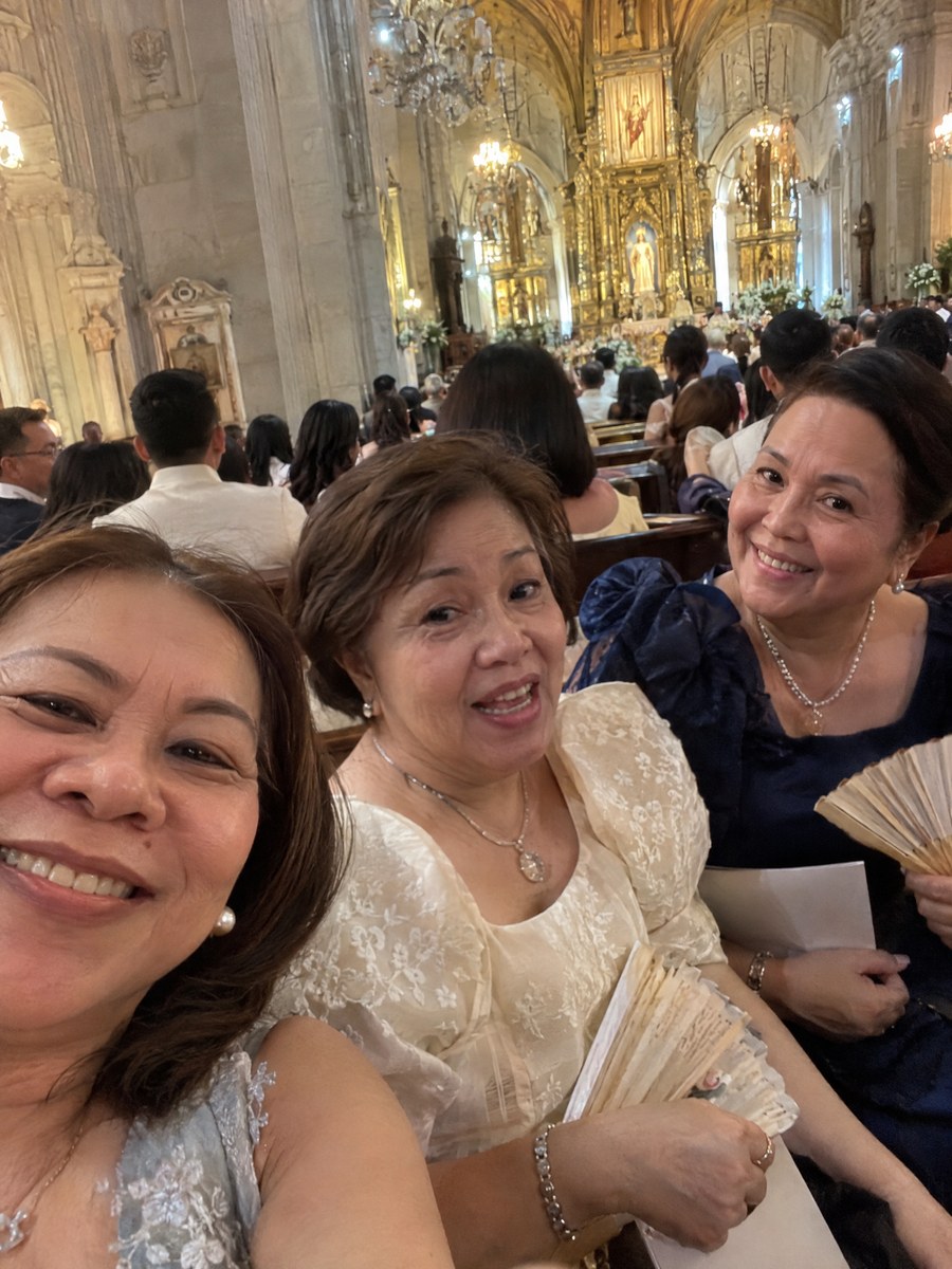 Three aunts squeezed into a phone selfie in a church pew, fans in hand.