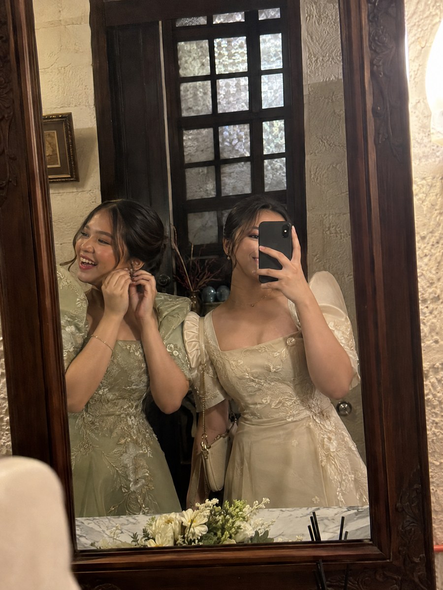 Two cousins in Filipiniana sleeves taking a mirror selfie in an old powder room.