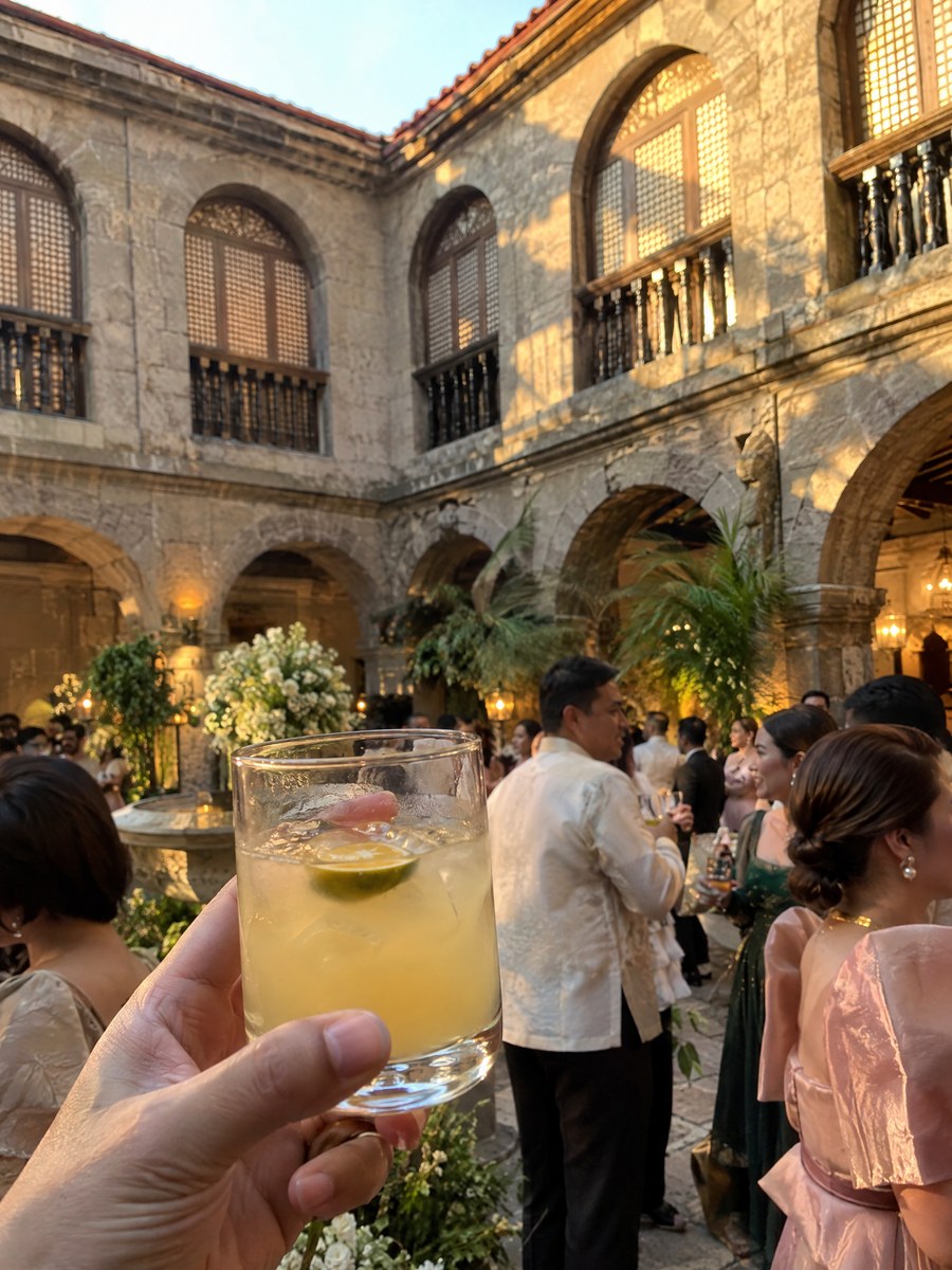 A glass of calamansi juice held up in the adobe courtyard at golden hour.