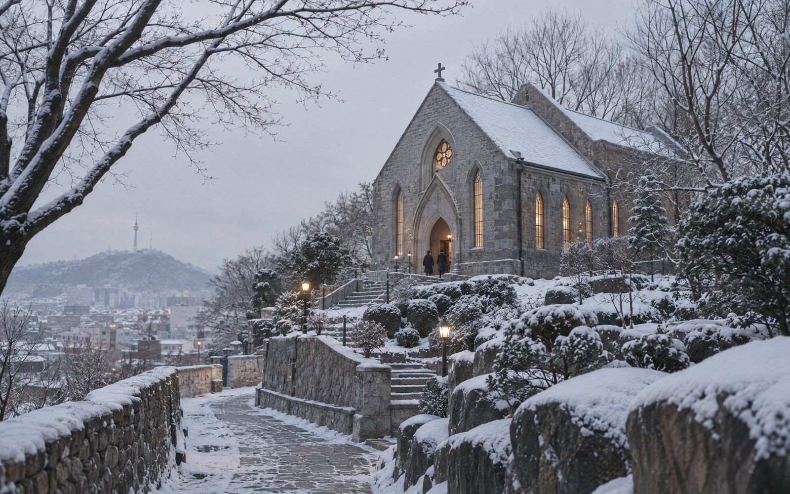 A small grey-stone chapel on a hillside with a snow-lined slate roof and warm light in its tall windows.