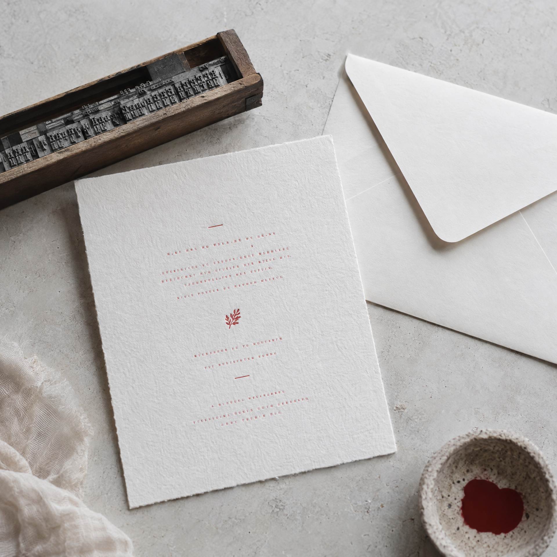 The invitation: a heavy white card with a deep letterpress impression in red ink, its envelope and a composing stick of metal type beside it.