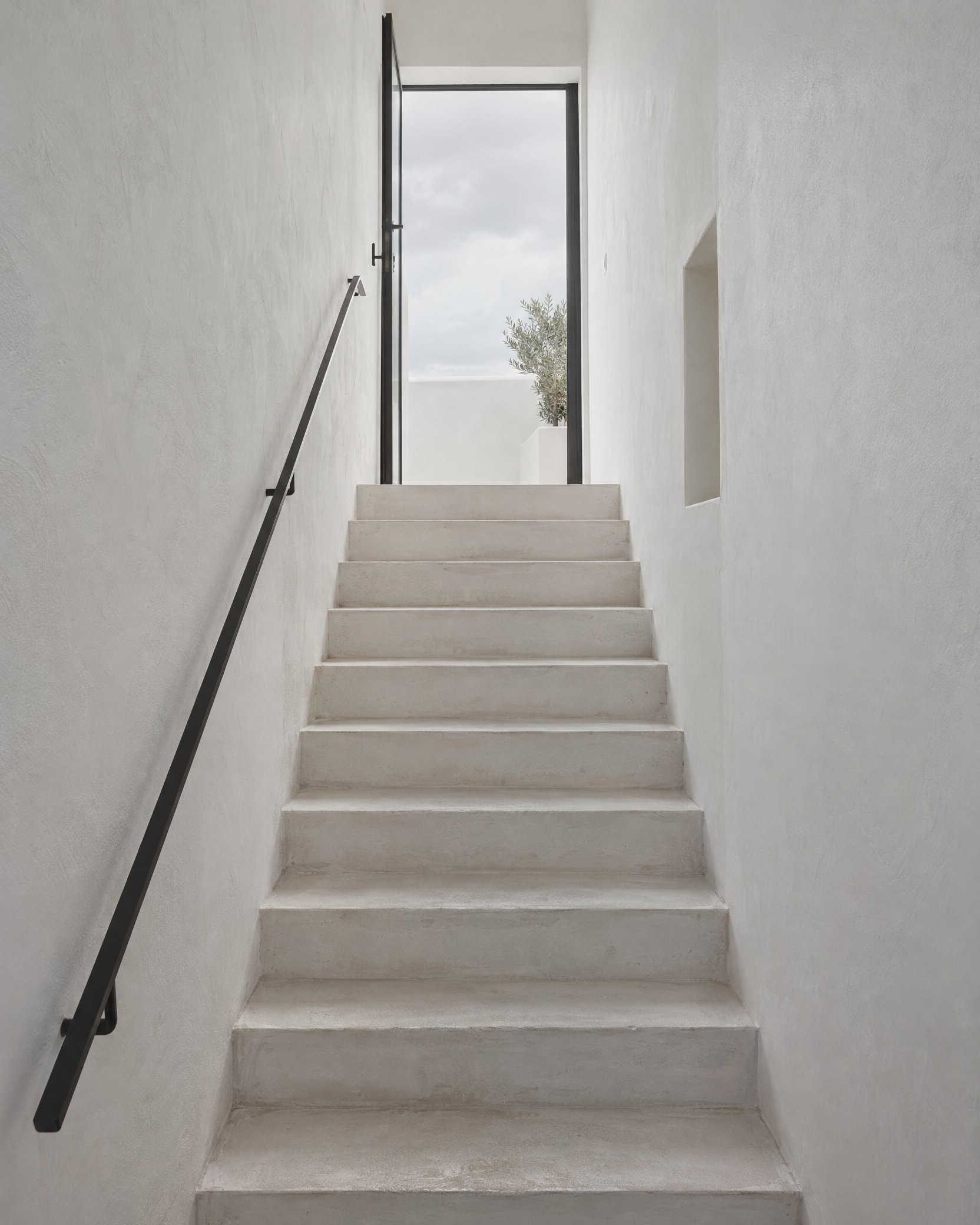 A narrow whitewashed stair with a thin black handrail, daylight falling in from the door at the top.